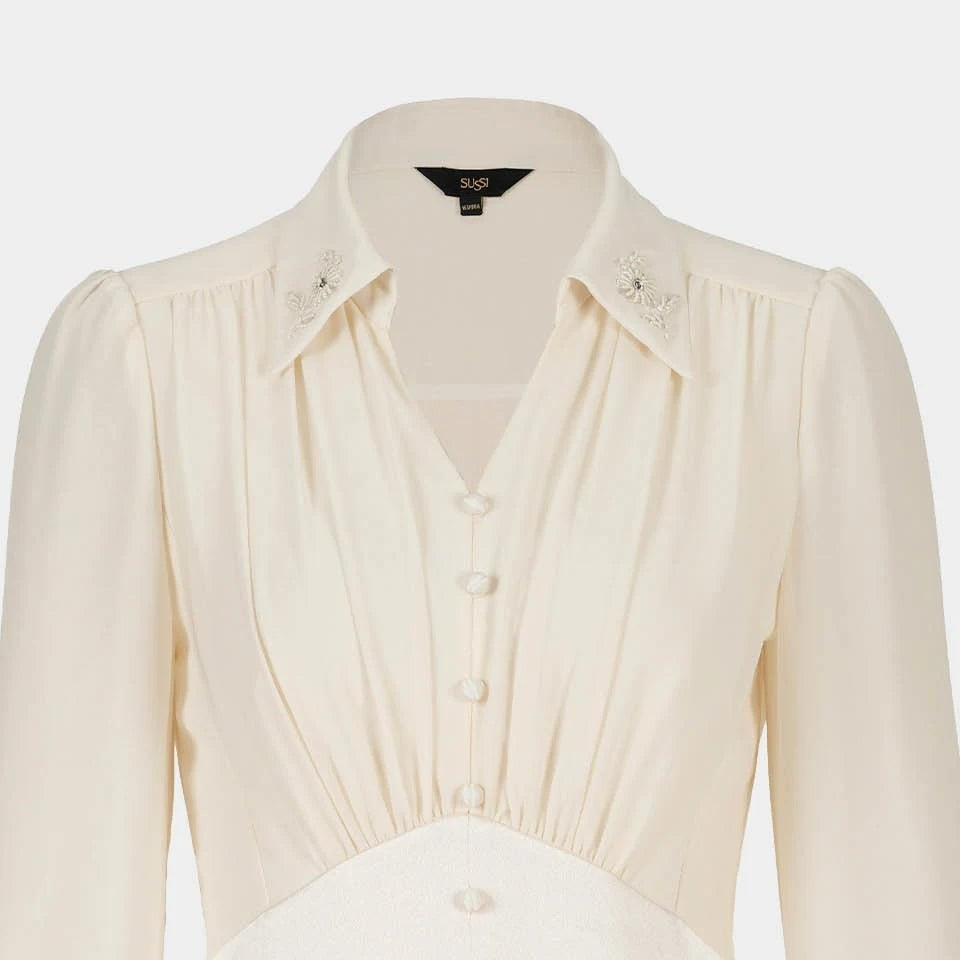 Narrow folds descend beneath the blouse's pointed collar towards a fitted waist section.